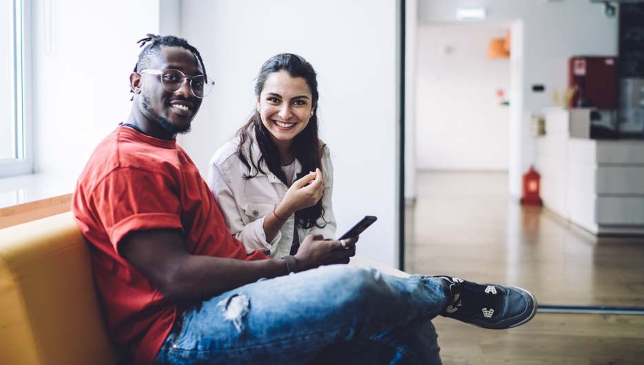 Side view of smiling casual black man with charming female group mate sitting and using smartphone while looking at camera
