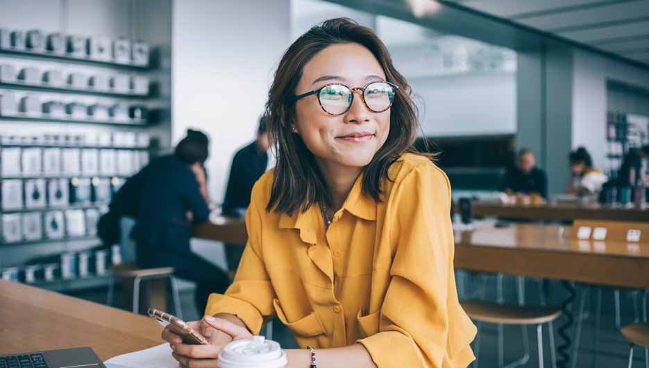 Portrait of cheerful hipster girl in optical spectacles for provide eyes protection looking at camera and smiling during blogging time in university classroom, concept of browsing and networking