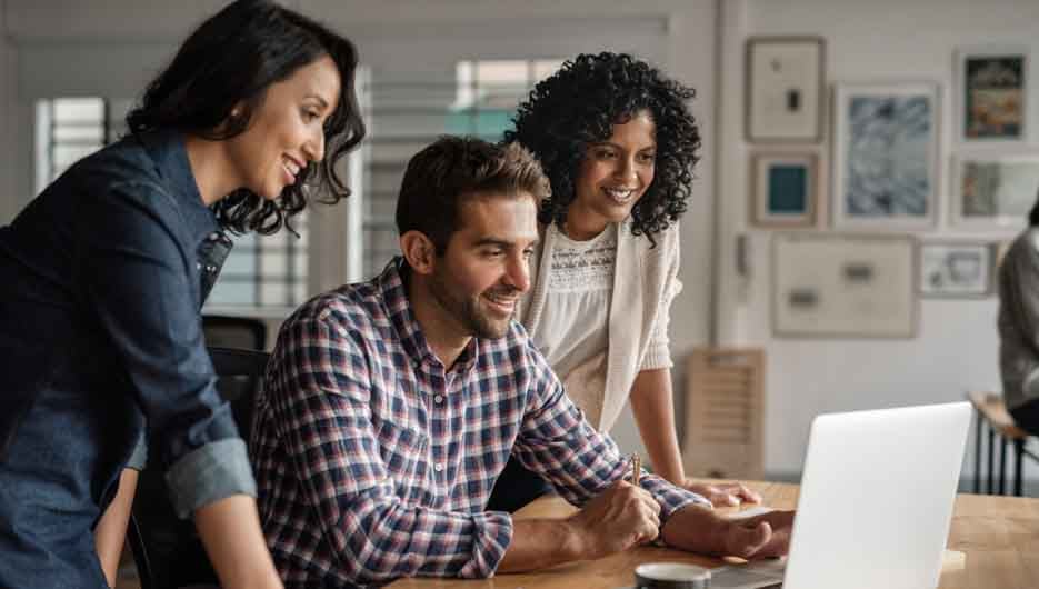 Smiling group of diverse young designers working on a laptop together at a desk in their startup office