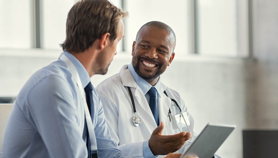Two mature smiling doctors having discussion about patient diagnosis, holding digital tablet. Successful african surgeon discussing case after positive result. Representative pharmaceutical discussing with african happy doctor about new medicine.