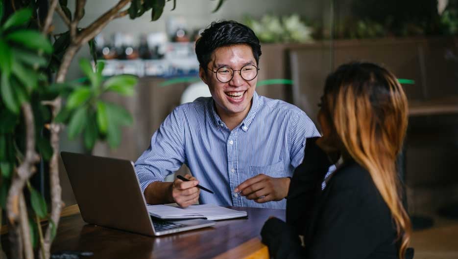 A confident and handsome young man is interviewing a female candidate for a job. They are sitting together in a table at a trendy coworking space. He is laughing and having a relaxed conversation.