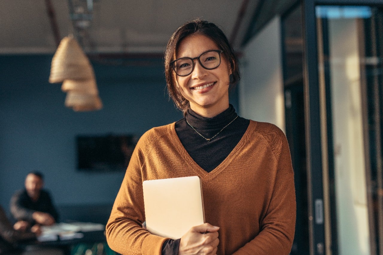 Portrait of happy business woman holding a laptop standing in office with colleagues working in background. Smiling woman in casuals in office.