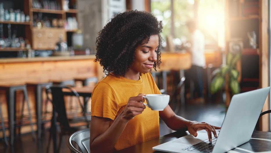 Portrait of an african smiling young woman sitting in cafe holding coffee cup in hand typing on laptop
