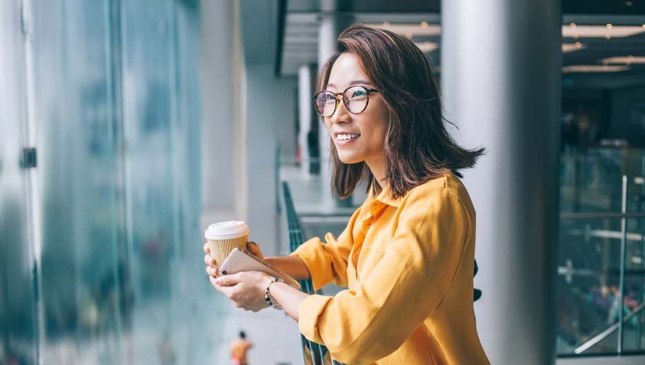 Side view of cheerful positive pretty Asian female in glasses and yellow shirt with hot drink and mobile phone leaning on handrail looking away and thinking in Hong Kong indoors